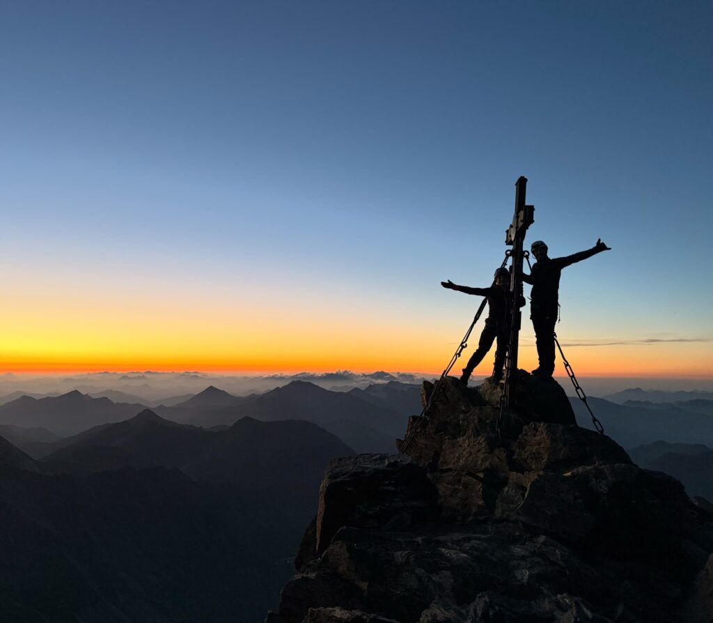 Zwei Menschen stehen bei Sonnenaufgang mit ausgestreckten Armen neben einem Kreuz auf einem Berggipfel, ihre Silhouetten heben sich vor einem farbenprächtigen Himmel ab, in dessen Ferne Bergketten zu sehen sind – ein unvergesslicher Moment auf dem Großglockner-Normalweg.