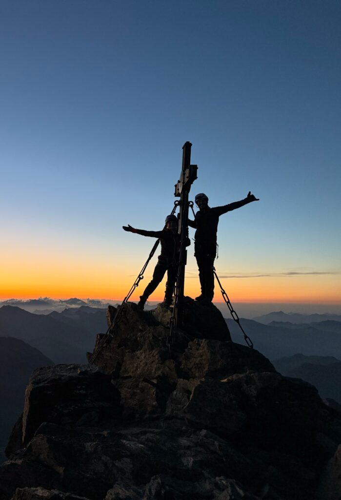 Zwei Menschen, die bei Sonnenuntergang mit ausgestreckten Armen neben einem Kreuz auf einem Berggipfel stehen, vor dem Hintergrund eines farbenprächtigen Himmels und ferner Bergketten – ein atemberaubender Moment, den man oft bei einer geführten Besteigung auf dem Großglockner-Normalweg erlebt.