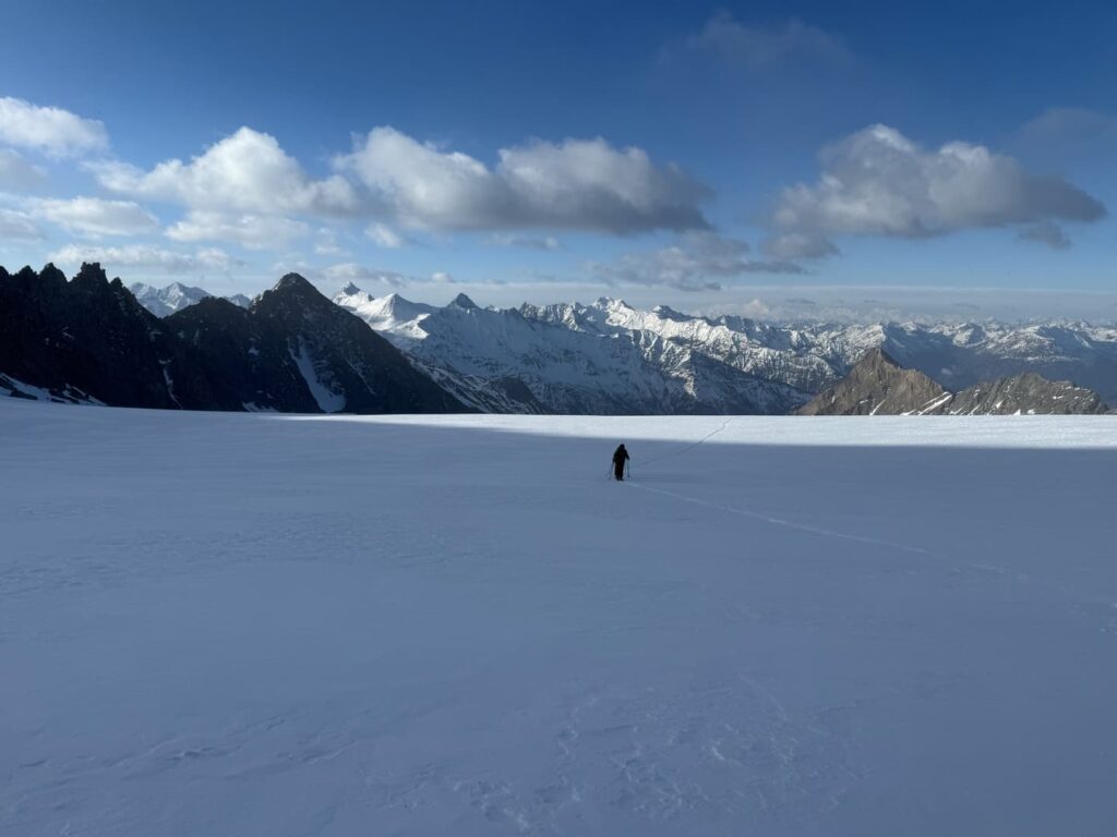 Eine einzelne Person wandert über ein weites, verschneites Feld, während sich in der Ferne unter einem teilweise bewölkten blauen Himmel schroffe, schneebedeckte Berge abzeichnen – ein Bild, das das atemberaubende Abenteuer einer geführten Skitour auf den Großglockner suggeriert.