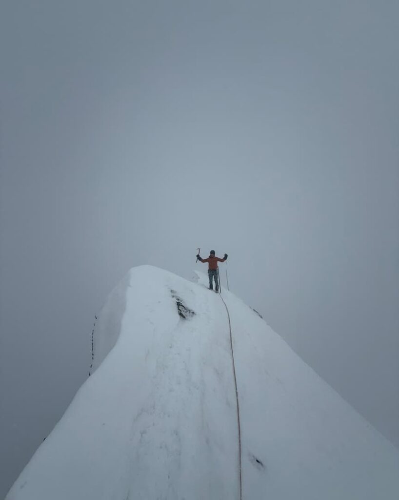 Ein Bergsteiger in einer roten Jacke steht triumphierend auf einem schneebedeckten Berggipfel und reckt beide Arme in die Höhe, während ein Kletterseil vor dem Hintergrund eines grauen, bewölkten Himmels hinter ihm herweht – ein atemberaubender Moment aus einer geführten Skitour auf den Großglockner.