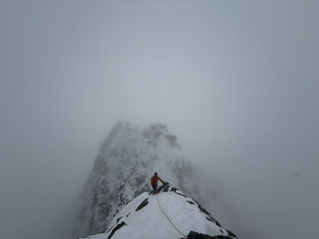 Ein Bergsteiger in einer roten Jacke erklimmt auf einer geführten Skitour am Großglockner einen schmalen, verschneiten Berggrat, der von dichtem Nebel umhüllt ist; vor ihm ist ein steiler, felsiger Gipfel zu sehen.