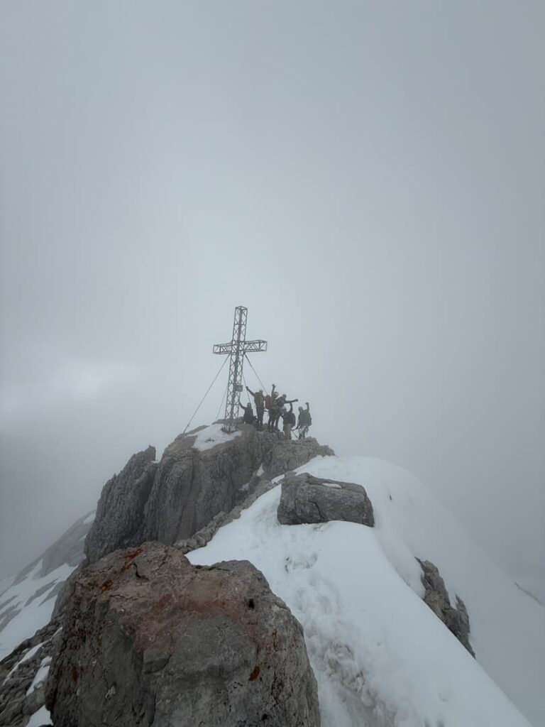 Eine Gruppe von Menschen steht auf einem schneebedeckten Berggipfel neben einem großen Metallkreuz, umgeben von Wolken und felsigem Gelände, nachdem sie gerade den Normalweg zum Hohen Dachstein bewältigt haben.