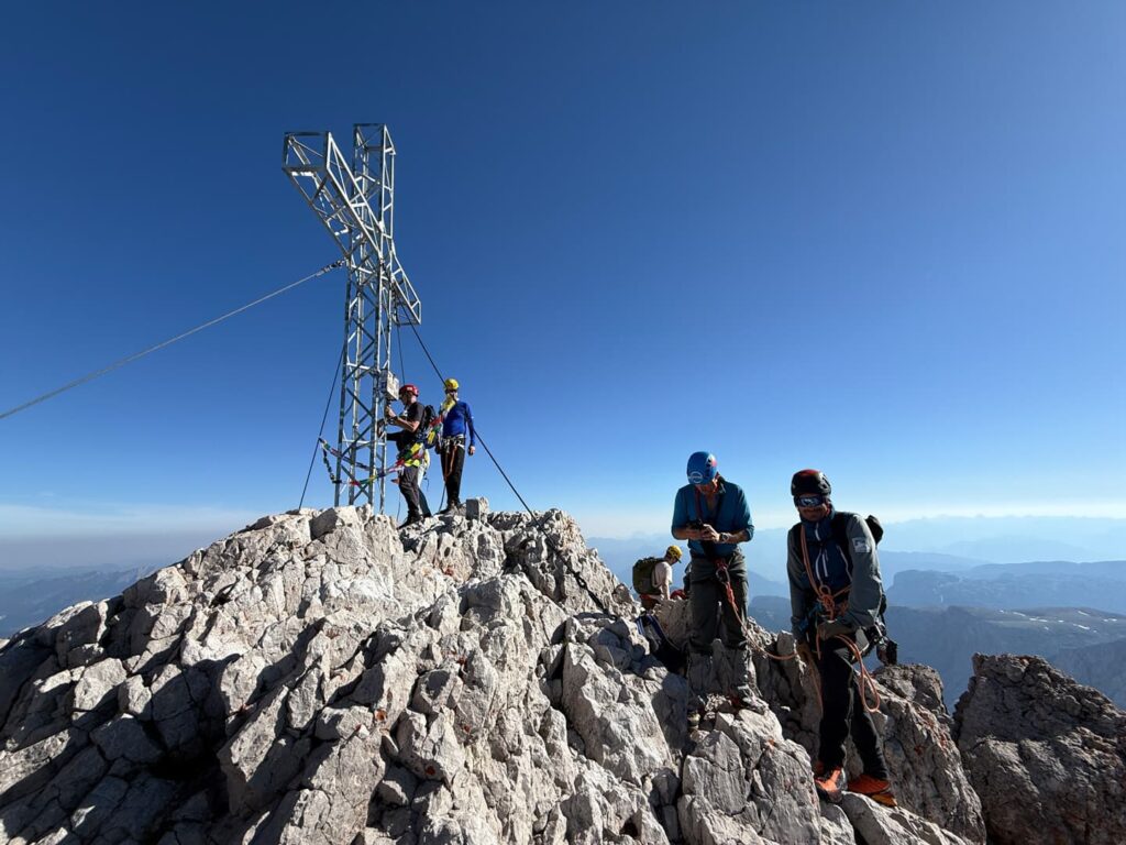 Mehrere Bergsteiger mit Helmen und Ausrüstung stehen auf dem Gipfel eines felsigen Berges in der Nähe eines großen Metallkreuzes; sie haben vermutlich den Normalweg zum Hohen Dachstein bewältigt, unter einem strahlend blauen Himmel, im Hintergrund sind ferne Berge zu sehen.
