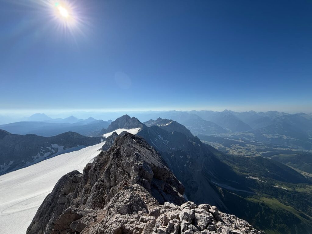 Ein felsiger Gebirgskamm unter strahlendem Sonnenschein, links davon ein schneebedeckter Abschnitt, mit Blick auf ferne Gipfel und einen strahlend blauen Himmel – diese Szene erinnert an die atemberaubenden Ausblicke entlang des Hoher Dachstein Normalwegs.