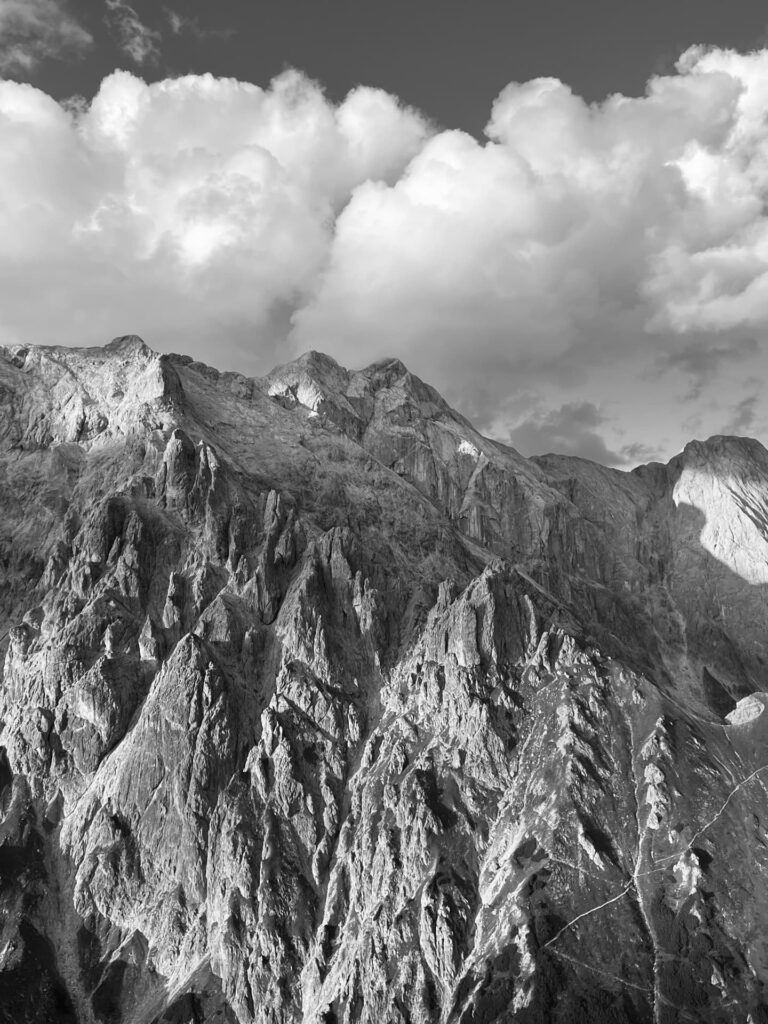 Im Vordergrund ragen schroffe Berggipfel steil empor, unter einem Himmel voller großer, wogender Wolken. Das Schwarz-Weiß-Bild hebt die Struktur der felsigen Hänge und die dramatische Lichtstimmung hervor – und fängt damit perfekt das anspruchsvolle Gelände ein, das auf einer „Königsjodler geführt“-Tour durchquert wird.