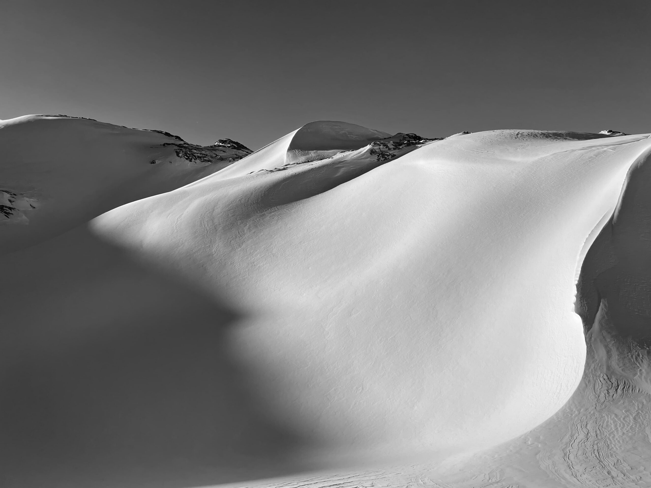 Schneebedeckte Berghänge mit sanften, wellenförmigen Konturen und tiefen Schatten unter klarem Himmel schaffen eine dramatische und doch ruhige Winterlandschaft in Schwarz-Weiß – das perfekte Terrain für geführte Bergtouren, die dazu einladen, diese atemberaubende Szenerie zu erkunden.