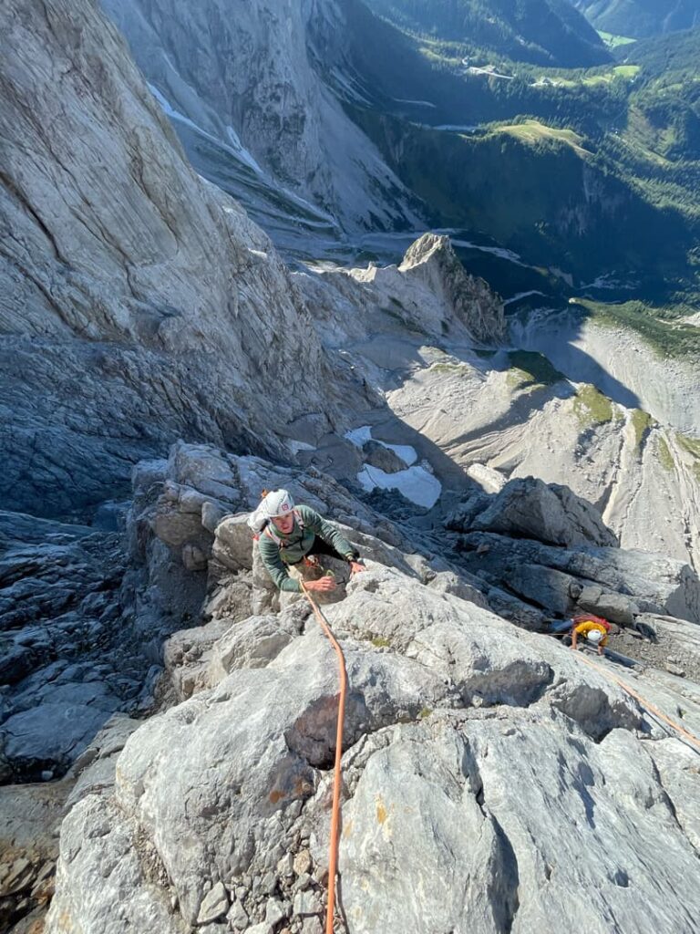 Ein Kletterer erklimmt eine steile, felsige Bergwand auf der berühmten Route „Pichlweg Dachstein geführt“ und ist dabei mit einem orangefarbenen Seil gesichert. Ein weiterer Kletterer folgt ihm darunter. Im Hintergrund sind dramatische Felswände und ein grünes Tal zu sehen.