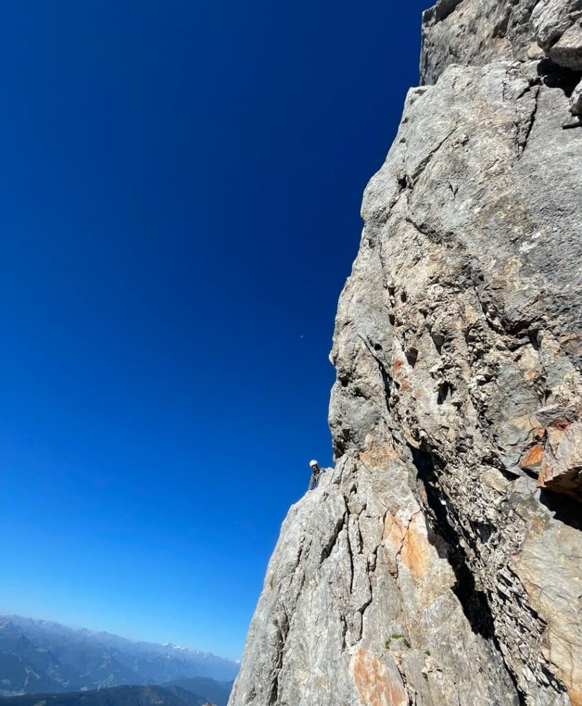 Ein Kletterer mit Helm erklimmt unter strahlend blauem Himmel eine steile, felsige Bergwand auf dem Steinerweg am Dachstein, während in der Ferne die Berge unter ihm zu sehen sind.