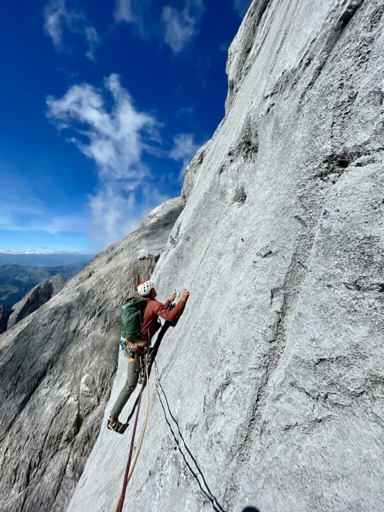 Ein Kletterer mit Helm und Ausrüstung erklimmt an einem sonnigen Tag eine steile, graue Felswand, während im Hintergrund der blaue Himmel und die Berge zu sehen sind, und genießt den Nervenkitzel eines „Königsjodler geführt“-Abenteuers.