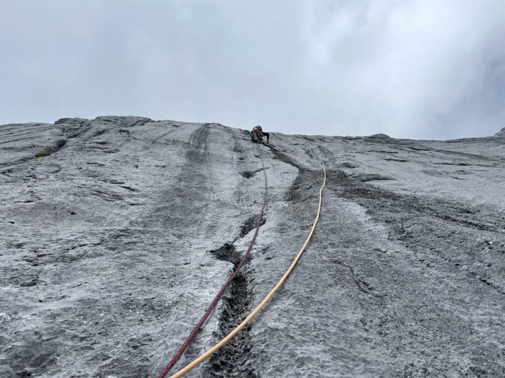 Ein Kletterer erklimmt eine steile, graue Felswand auf der Route „Königsjodler geführt“ und ist dabei durch Seile gesichert, die unter dem bewölkten Himmel in Richtung Kamera hinabreichen.