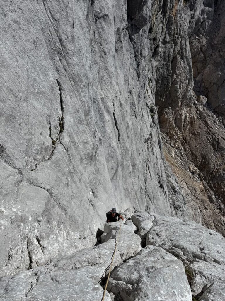 Ein Kletterer erklimmt mithilfe eines Seils eine steile, graue Felswand, umgeben von schroffen Steinwänden und zerklüfteten Formationen in einer bergigen Landschaft – eine atemberaubende Szene, die an die legendären Routen des Steinerwegs am Dachstein erinnert.