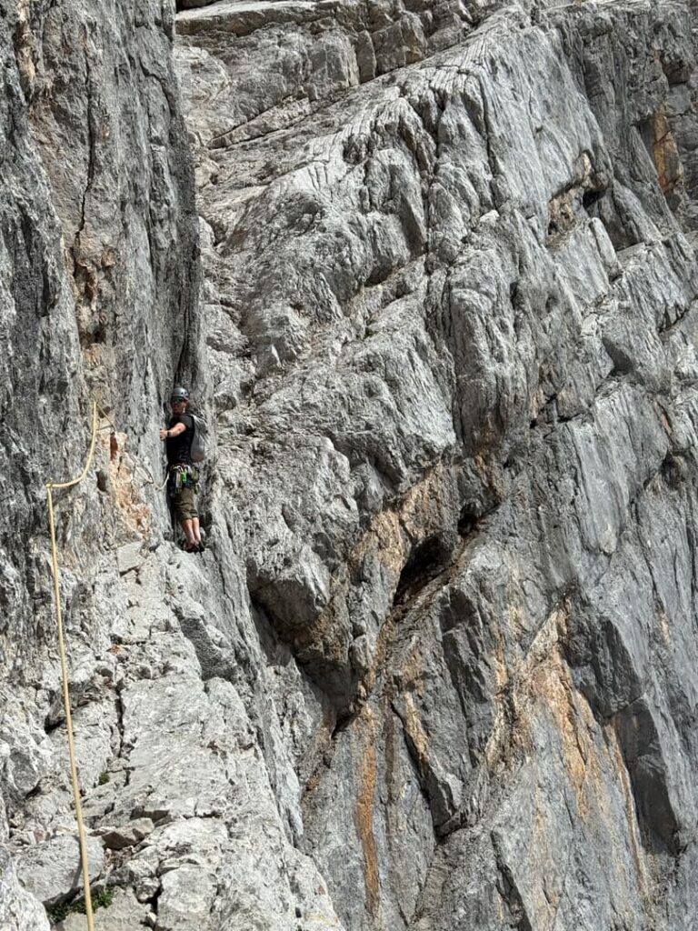 Eine Person mit Helm und Kletterausrüstung erklimmt eine steile, zerklüftete Felswand, gesichert durch ein Seil – ein Erlebnis, das an die legendären „Steinerweg Dachstein geführt“-Routen erinnert. Das felsige Gelände umgibt sie und unterstreicht die Größe und die Herausforderung des Aufstiegs.