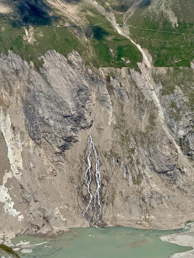 Ein hoher, schmaler Wasserfall stürzt einen felsigen Berghang hinab in einen blass türkisfarbenen Fluss, umgeben von schroffer Landschaft und grünen Grasflächen am oberen Rand – was an die dramatische Kulisse entlang des Großglockner-Normalwegs erinnert.