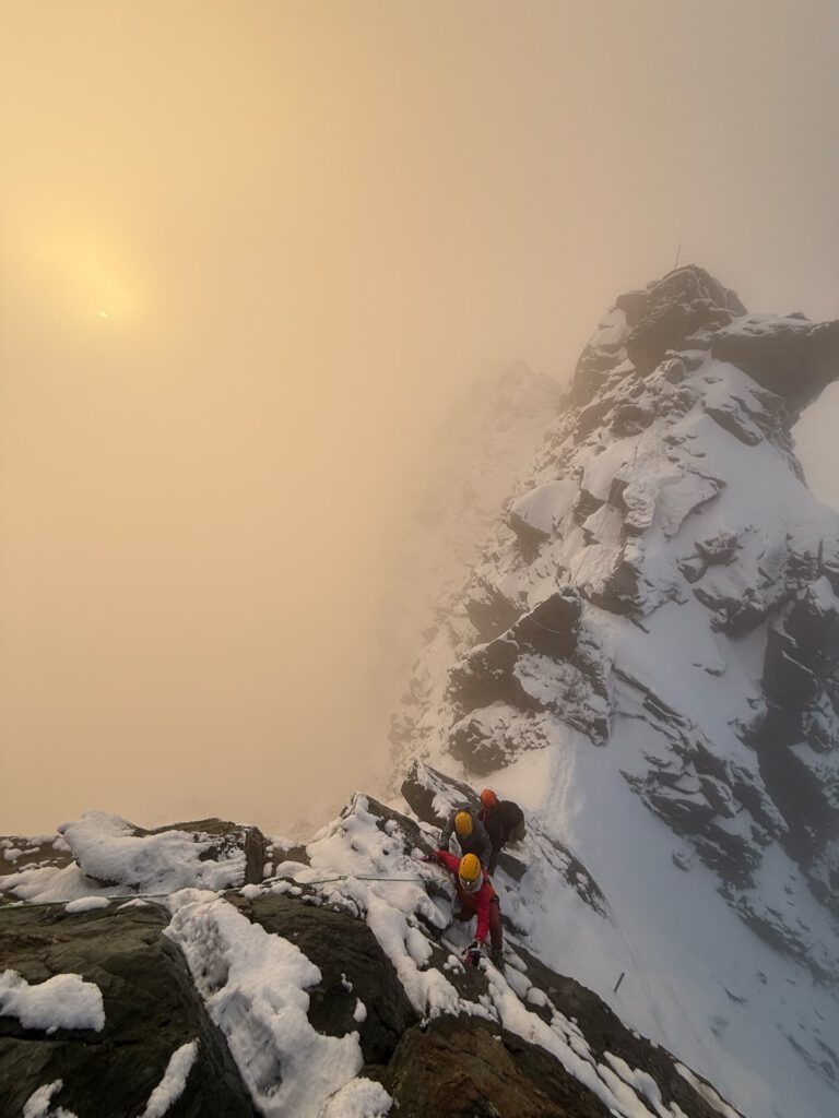 Zwei Bergsteiger mit Helmen steigen bei Sonnenaufgang oder Sonnenuntergang einen verschneiten, felsigen Berggrat entlang des Großglockner-Normalwegs hinauf, vor dem Hintergrund eines nebligen, goldenen Himmels.