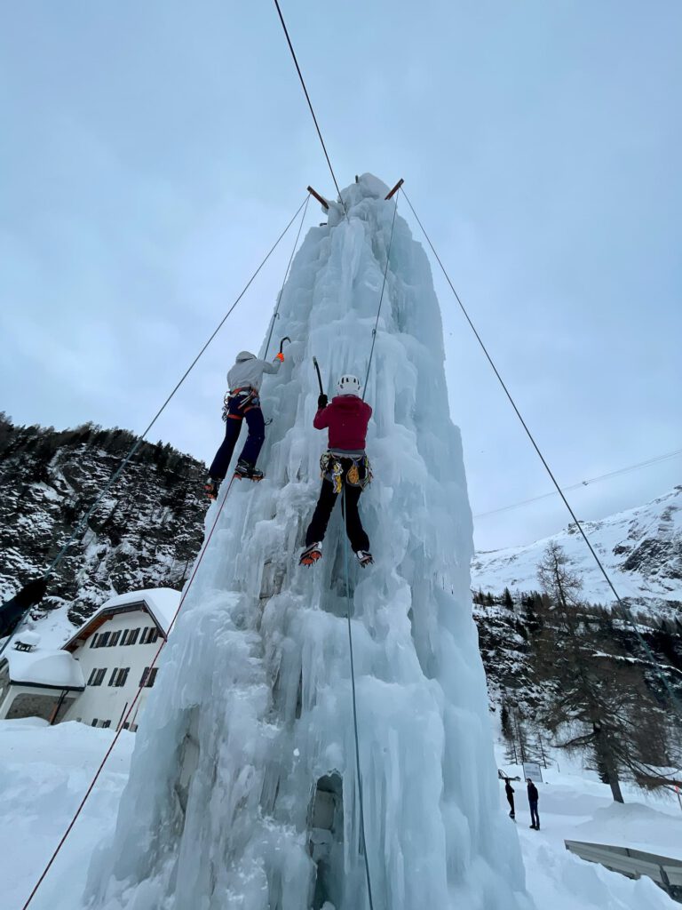 Zwei Personen mit Helmen und Winterausrüstung klettern im Rahmen eines Eiskletterkurses in den Hohen Tauern im Freien an einer hohen, massiven Eissäule entlang. Dabei verwenden sie Seile und Kletterausrüstung, während im Hintergrund schneebedeckte Berge und Bäume zu sehen sind.