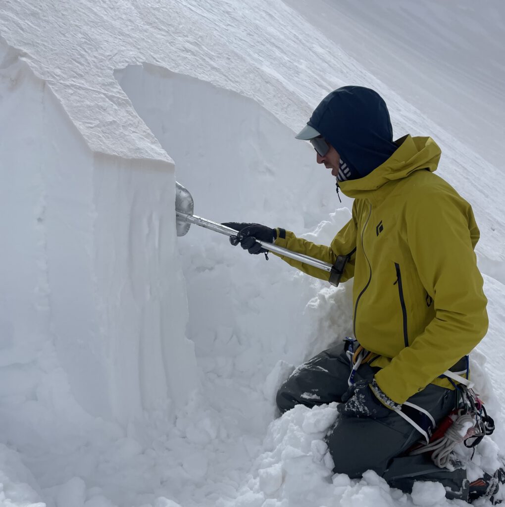 Eine Person in einer gelben Jacke untersucht mit einer Schaufel eine Schneewand, wahrscheinlich um die Schneeschichten oder die Stabilität in einer verschneiten Berglandschaft zu prüfen – eine wichtige Sicherheitsmaßnahme, die häufig bei geführten Bergtouren durchgeführt wird.