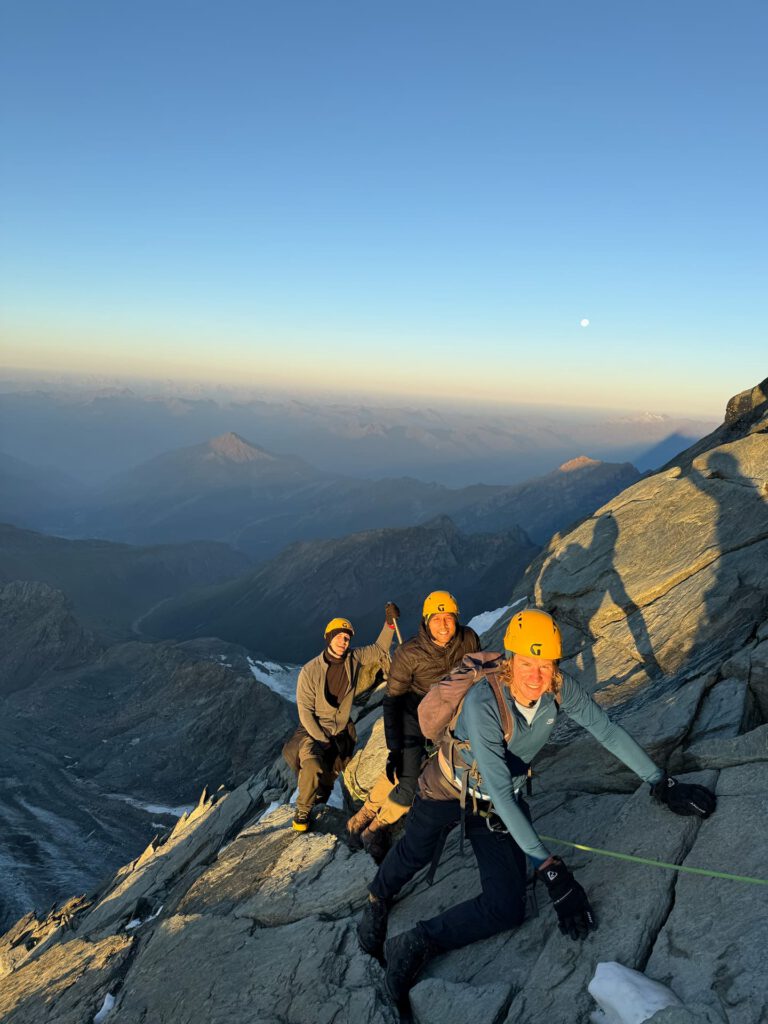 Drei Bergsteiger mit Helmen und Klettergurten erklimmen bei Sonnenaufgang auf dem Großglockner-Normalweg einen steilen, felsigen Berghang, im Hintergrund erstreckt sich eine weite Berglandschaft und in der Ferne ist der Mond zu sehen.
