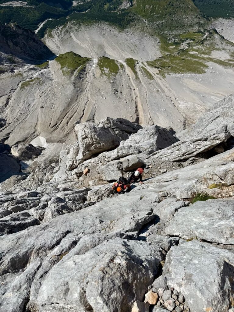 Zwei Bergsteiger mit Helmen erklimmen einen steilen, felsigen Berghang entlang der Route „Pichlweg Dachstein geführt“, während im Hintergrund weit unter ihnen unwegsames Gelände und grüne Flecken zu sehen sind.