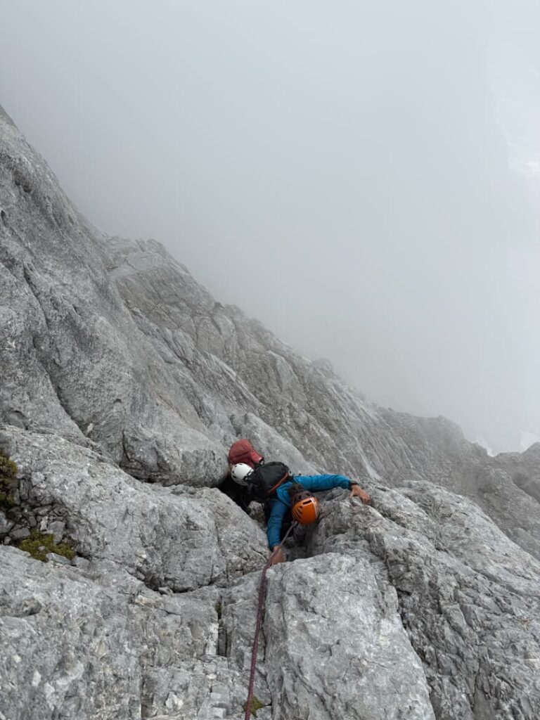 Zwei Bergsteiger mit Helmen erklimmen eine steile, felsige Bergwand, die von Nebel umhüllt ist, und lassen sich dabei von Seilen sichern, während sie die berühmte Route „Pichlweg Dachstein geführt“ hinaufsteigen.