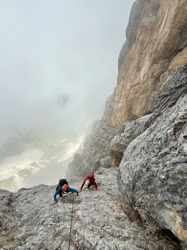 Zwei Kletterer mit Helmen und Ausrüstung erklimmen die steile, felsige Bergwand der Route „Pichlweg Dachstein geführt“, die teilweise von Nebel und Wolken umgeben ist, mit einem dramatischen Abgrund zur Linken und schroffen Felsformationen rundherum.