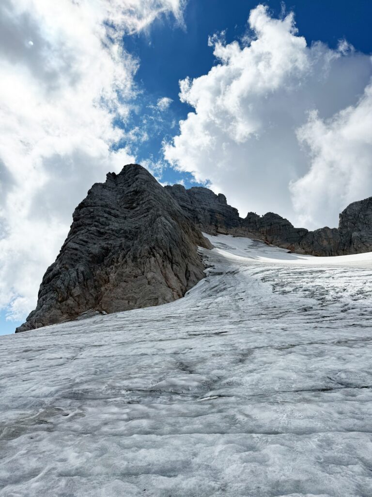 Ein zerklüfteter Felsgipfel ragt über einen schneebedeckten Hang unter einem strahlend blauen Himmel mit vereinzelten Wolken empor, entlang der klassischen Route „Hoher Dachstein Normalweg“.