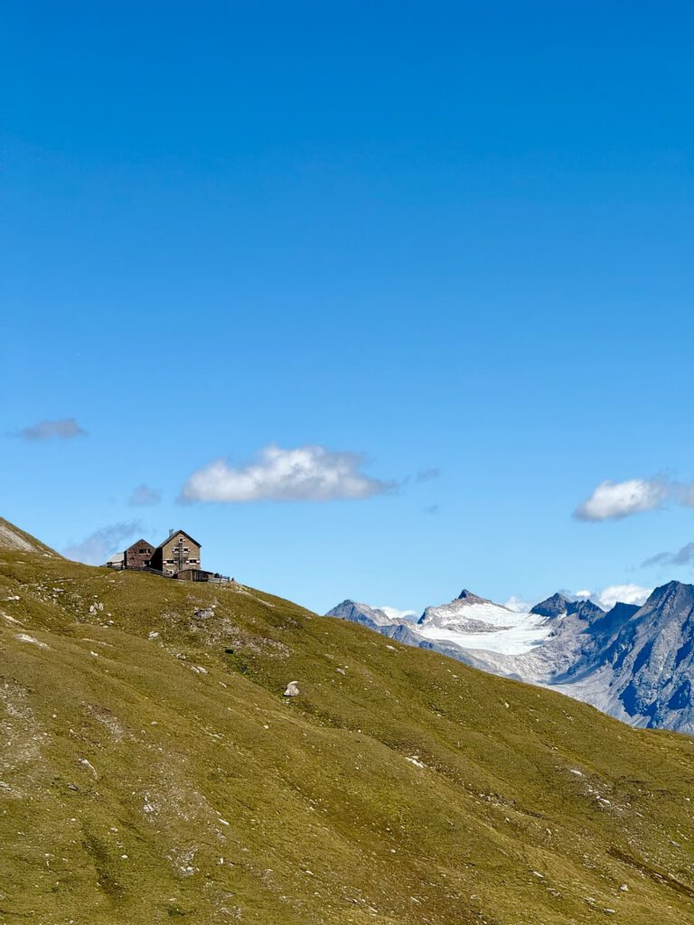 Unter einem strahlend blauen Himmel steht ein kleines Haus auf einem grasbewachsenen Hang, im Hintergrund sind in der Ferne schneebedeckte Berge – darunter die berühmte Großglockner-Normalweg-Route – und ein paar Wolken zu sehen.