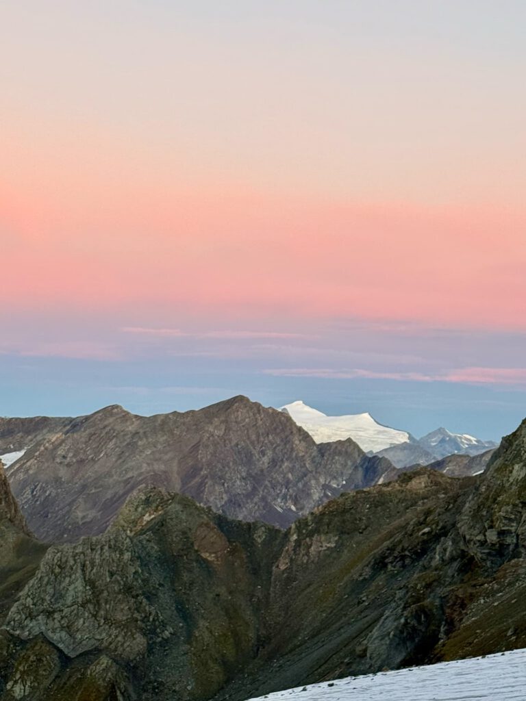 Schneebedeckte Berge unter einem rosa-blauen Abendhimmel, mit felsigen Gipfeln im Vordergrund und einem weißen Gipfel in der Ferne, der in sanftes Licht getaucht ist – ein unvergesslicher Anblick vom legendären Stüdlgrat-Aufstieg zum Großglockner.