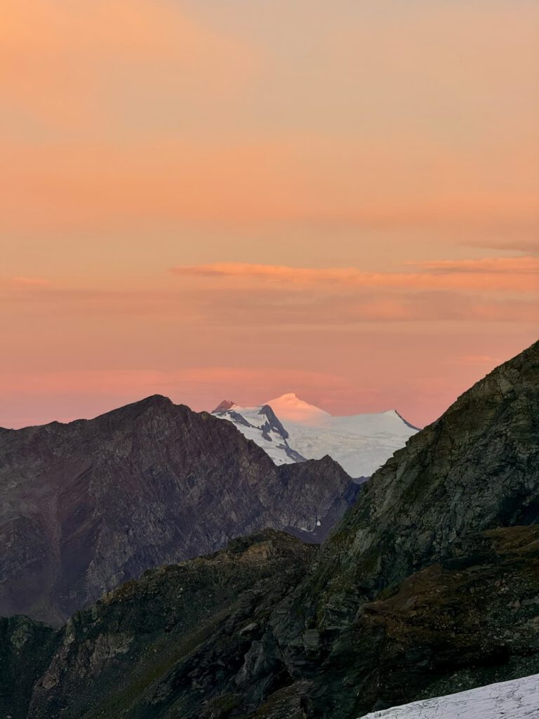 Ein schneebedeckter Berggipfel leuchtet bei Sonnenuntergang orange, eingerahmt von dunkleren Felsbergen im Vordergrund unter einem rosa-orangefarbenen Himmel mit vereinzelten Wolken – ein atemberaubender Anblick, der an das alpine Erlebnis „Stüdlgrat-Großglockner-Tour“ erinnert.