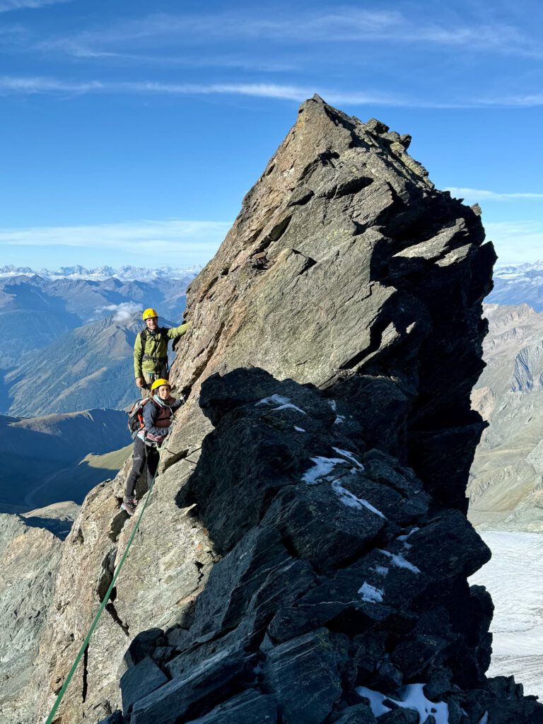 Zwei Bergsteiger mit Helmen erklimmen im Rahmen einer geführten Tour unter strahlend blauem Himmel den steilen, felsigen Stüdlgrat am Großglockner, umgeben von fernen Gipfeln und Tälern.