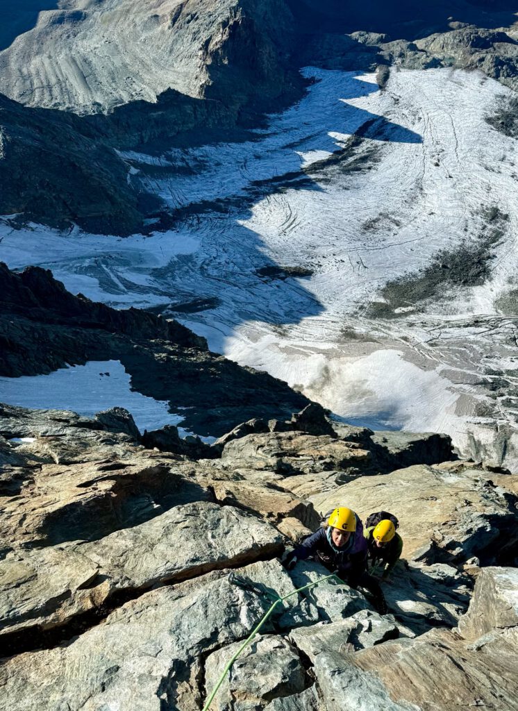 Zwei Bergsteiger mit gelben Helmen erklimmen einen steilen, felsigen Berghang auf der legendären Stüdlgrat-Großglockner-Führungsroute, gesichert durch Seile, während weit unter ihnen ein riesiger Gletscher und schroffe Gipfel zu sehen sind.