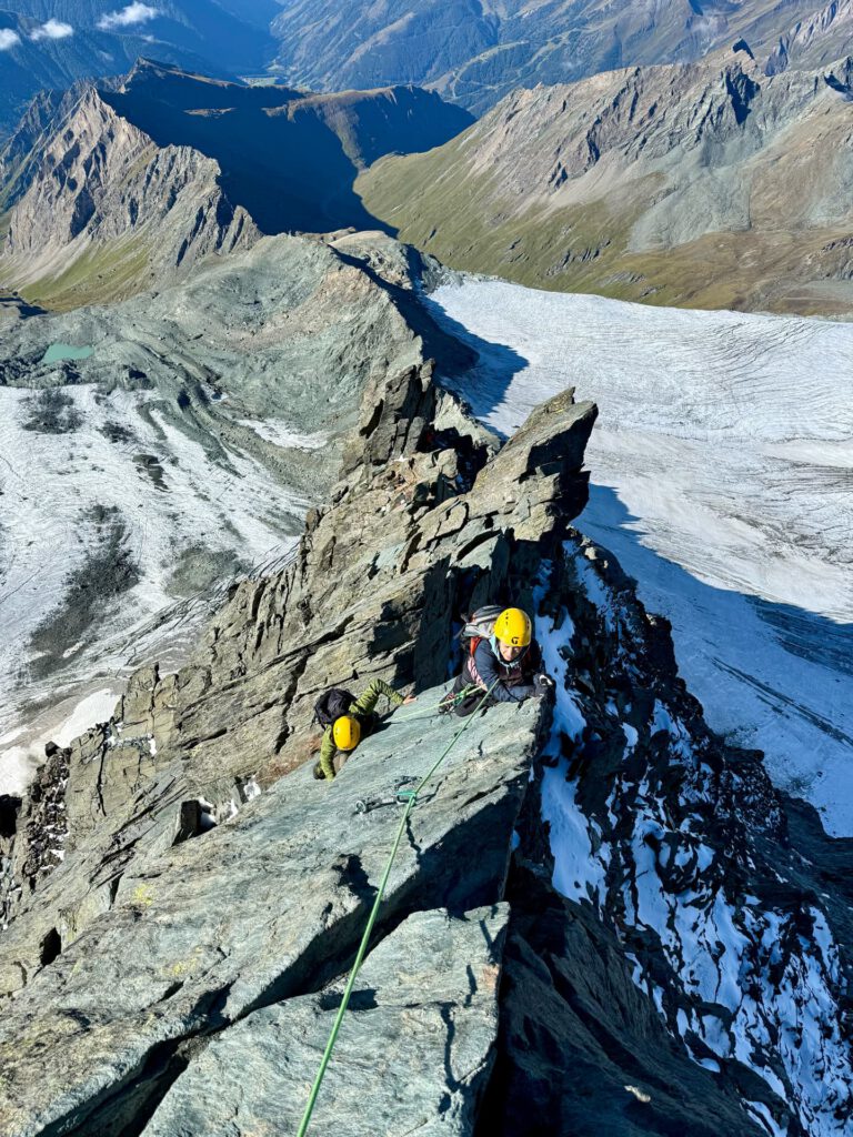 Zwei Bergsteiger mit gelben Helmen erklimmen den schmalen, zerklüfteten Stüdlgrat des Großglockners, gesichert durch ein Seil, während unter ihnen bei klarem Himmel ein Gletscher und schroffe Gipfel zu sehen sind – ein klassisches „geführtes“ alpines Abenteuer.