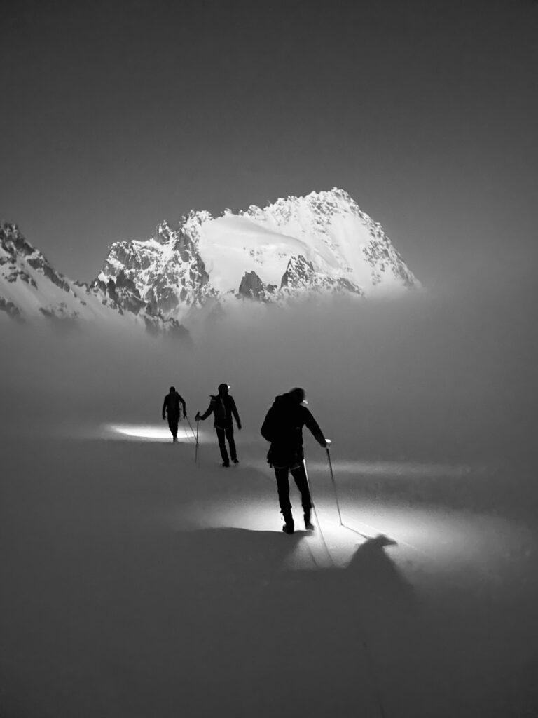 Drei Menschen wandern nachts durch den Schnee und beleuchten ihren Weg mit Stirnlampen – eine eindrucksvolle Szene, die den Geist geführter Bergtouren einfängt. Im Hintergrund ragen schroffe, schneebedeckte Berggipfel empor, die teilweise im Nebel verborgen sind. Die Szene ist in Schwarz-Weiß gehalten.