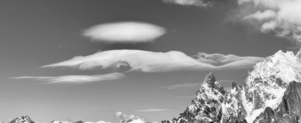 Schneebedeckte Berggipfel unter einem größtenteils klaren Himmel, über dem ungewöhnliche, glatte Linsenwolken schweben. Das von staatlich geprüften Bergführern aufgenommene Schwarz-Weiß-Bild bringt die dramatische Landschaft aus der Perspektive eines Experten zur Geltung.