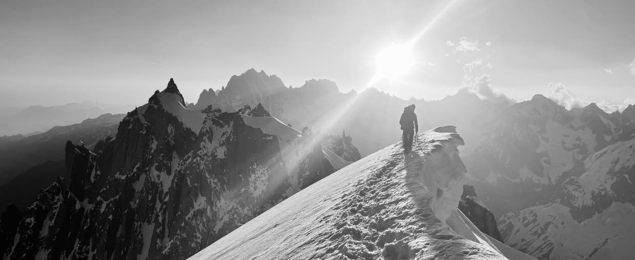 Ein einzelner Bergsteiger, begleitet von einem Bergführer von ARE YOU SUMMIT, wandert bei Sonnenaufgang über einen verschneiten Bergrücken, im Hintergrund ragen zerklüftete Gipfel empor und Sonnenstrahlen durchziehen den Himmel.