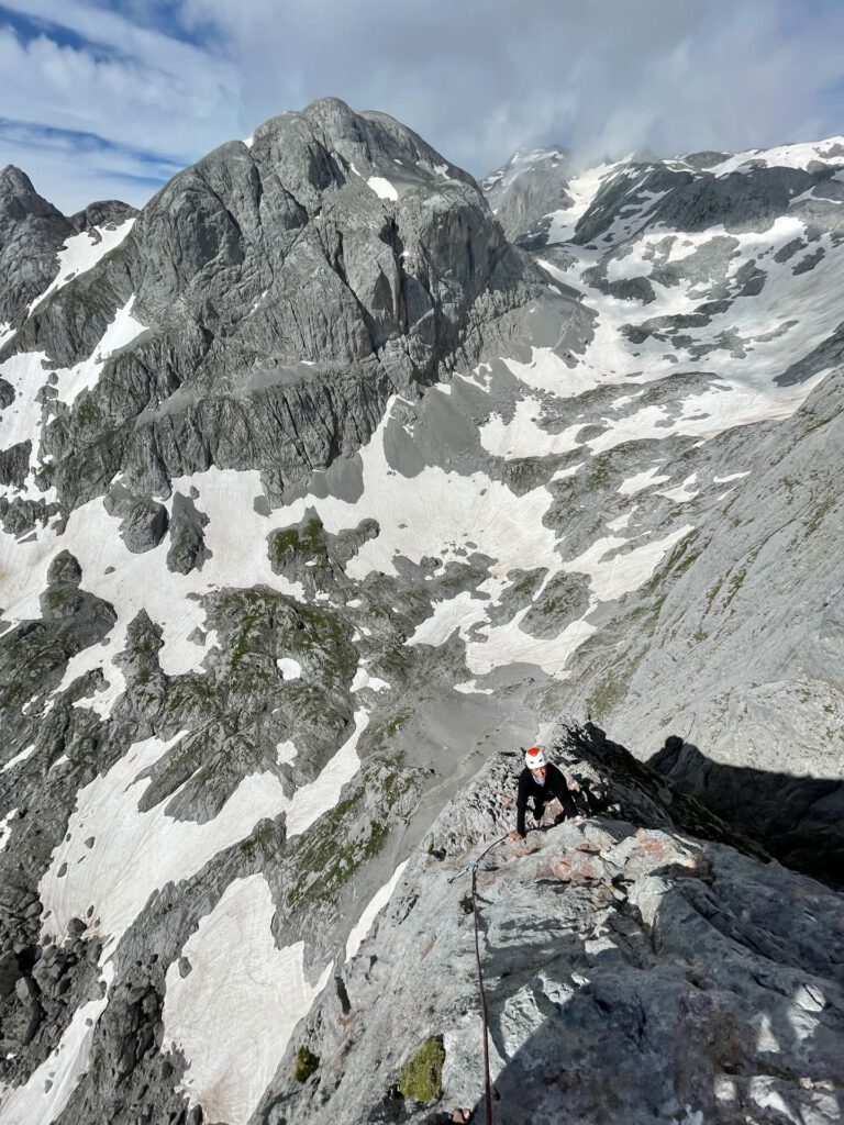 Ein Kletterer mit Helm erklimmt den felsigen Königsjodler-Grat, umgeben von steilen grauen Felsen und Schneeflecken unter einem teilweise bewölkten Himmel.