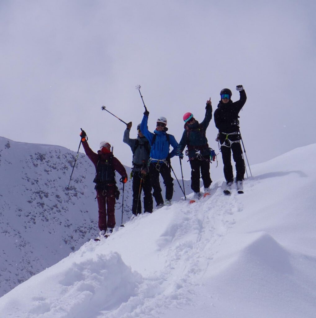 Fünf Menschen in Winterkleidung stehen auf einem verschneiten Berggipfel und recken jubelnd ihre Wanderstöcke in die Höhe. Der Himmel ist bewölkt, und Schnee bedeckt die umliegenden Hänge – eine perfekte Kulisse für geführte Bergtouren, die Abenteuer und Kameradschaft inmitten atemberaubender Alpenlandschaft versprechen.