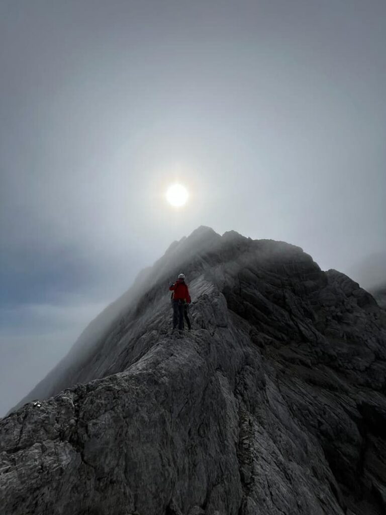 Eine Person in einer roten Jacke steht auf dem felsigen Berggrat des Steinerwegs am Dachstein unter einem dunstigen Himmel, während die Sonne durch den Nebel über den Gipfeln scheint.