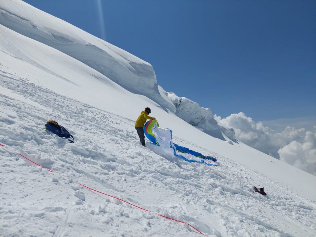 Eine Person in einer gelben Jacke bereitet unter strahlend blauem Himmel auf einem verschneiten Berghang ein farbenfrohes Gleitschirmsegel vor und macht sich bereit für ein aufregendes „Hike & Fly“-Abenteuer. In der Nähe liegen Skiausrüstung und Zubehör verstreut.