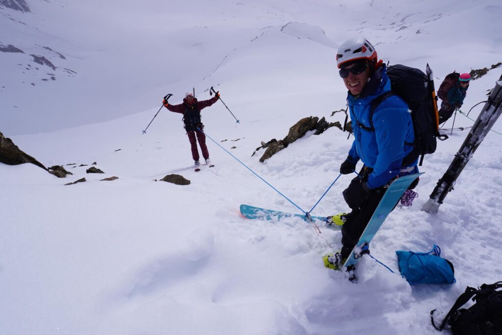Drei Personen in Skiausrüstung stehen während einer ihrer geführten Skihochtouren in den Alpen auf einem verschneiten Berghang; eine lächelt in die Kamera, während eine andere die Skistöcke in einer feierlichen Geste in die Höhe reckt. Um sie herum liegen Skier, Rucksäcke und Schnee.
