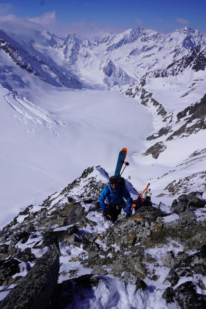 Eine Person in einer blauen Jacke erklimmt einen felsigen, verschneiten Berggrat und trägt dabei Skier – eine Szene, die symbolisch für geführte Skihochtouren in den Alpen steht. Im Hintergrund erstrecken sich unter klarem Himmel schneebedeckte Gipfel und ein weitläufiger Gletscher.
