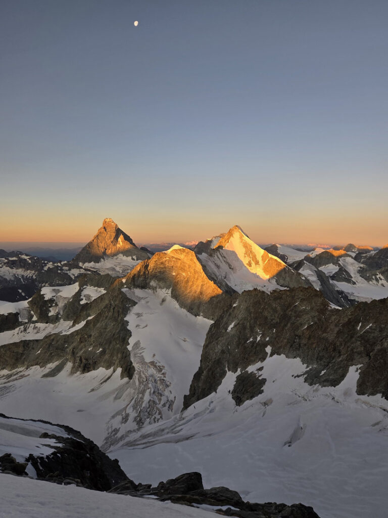 Schneebedeckte Berggipfel bei Sonnenaufgang, mit goldenem Licht auf den Gipfeln des Obergabelhorns und des Zinalrothorns. Der Himmel ist klar, darüber ist ein kleiner Mond zu sehen. Felsgrate und Gletscher prägen den Vorder- und Hintergrund und wecken Gedanken an geführte alpine Abenteuer.