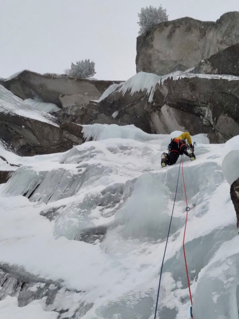 Ein Kletterer in Winterausrüstung erklimmt mithilfe von Seilen und Eispickeln eine vereiste, schneebedeckte Felswand und verkörpert damit die aufregende Herausforderung des Eiskletterns inmitten von Eiszapfen und Schnee auf den umliegenden Felsen.
