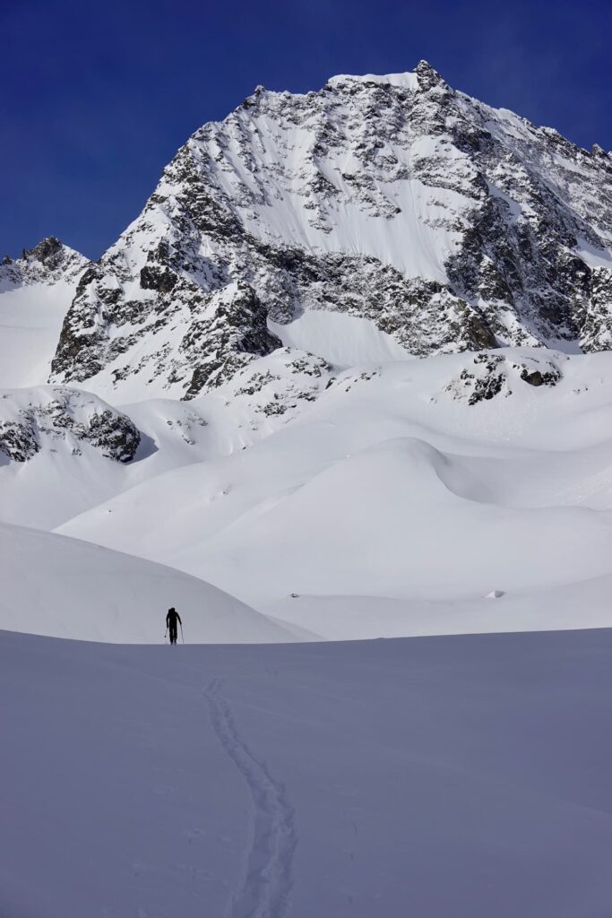 Eine einsame Gestalt wandert unter strahlend blauem Himmel durch tiefen Schnee auf einen hoch aufragenden, schroffen, schneebedeckten Berg zu und hinterlässt dabei eine einzige Spur aus Fußabdrücken – eine Szene, die an die Einsamkeit und das Abenteuer auf geführten Skitouren erinnert.