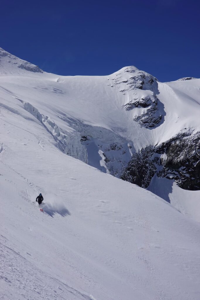 Ein Skifahrer fährt unter strahlend blauem Himmel einen steilen, verschneiten Berghang hinunter und hinterlässt dabei eine Spur im frischen Pulverschnee, während im Hintergrund schroffe Gipfel und vereiste Grate zu sehen sind – eine Szene, die an geführte Skidurchquerungen in den Alpen erinnert, bei denen Abenteurer unter fachkundiger Führung atemberaubende Alpenlandschaften durchqueren.