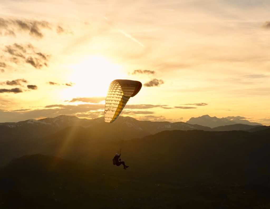 Eine Person genießt ein „Hike & Fly“-Abenteuer und gleitet bei Sonnenuntergang über dunkle Bergkämme, während die Sonne und goldene Wolken den Himmel im Hintergrund erhellen.