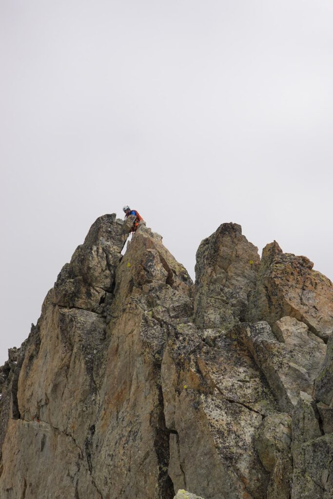 Ein Bergsteiger mit Helm und Rucksack erklimmt unter bewölktem Himmel zerklüftete, felsige Gipfel und verdeutlicht damit die Weite und die Herausforderungen des unwegsamen Berggeländes bei einer geführten Mont-Blanc-Tour.