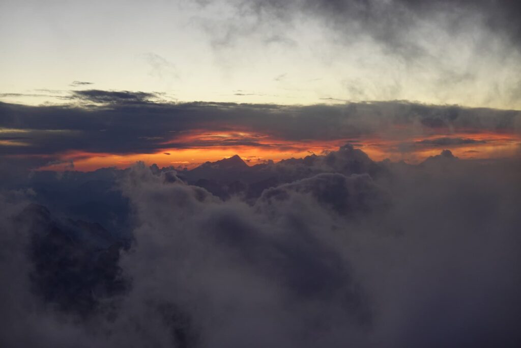 Wolken bedecken den Vordergrund, während in der Ferne zerklüftete Berggipfel zu sehen sind, die auf ein geführtes Abenteuer am Mont Blanc hindeuten. Ein leuchtend orange-gelber Sonnenuntergang strahlt am Horizont unter dramatischen dunklen Wolken.