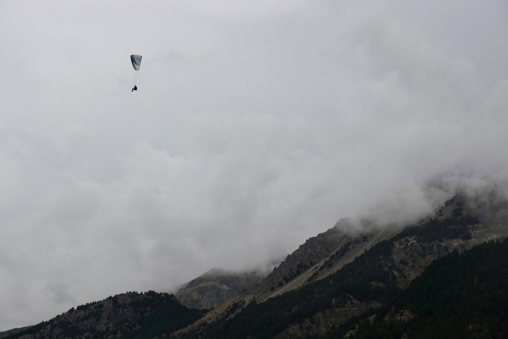 Ein einzelner Gleitschirmflieger schwebt unter einem bewölkten, grauen Himmel über neblige, bewaldete Berge und fängt so den Geist eines „Hike & Fly“-Abenteuers ein.