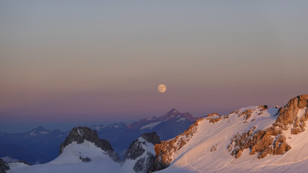 Bei Sonnenuntergang geht der Vollmond über den schneebedeckten Berggipfeln auf und taucht die Landschaft unter klarem Himmel in ein warmes, rosafarbenes Licht – eine bezaubernde Szene, die an ein geführtes Abenteuer am Mont Blanc erinnert.
