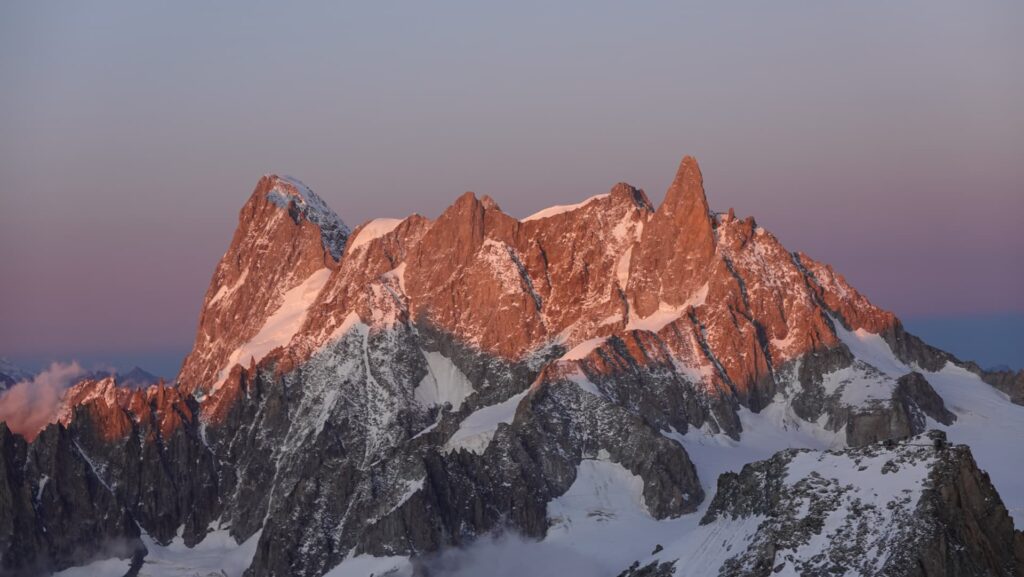 Schneebedeckte Berggipfel, wie beispielsweise bei einer geführten Tour auf den Mont Blanc, leuchten im sanften Licht des Sonnenaufgangs oder Sonnenuntergangs unter klarem Himmel in einem rosa-orangefarbenen Schimmer.